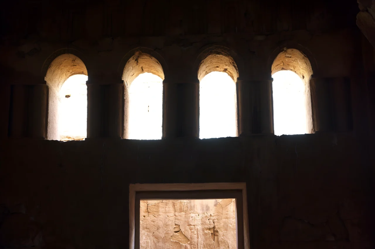 Quatre arches en plein cintre en pierre ocre ouvrant sur un ciel lumineux depuis une salle obscure